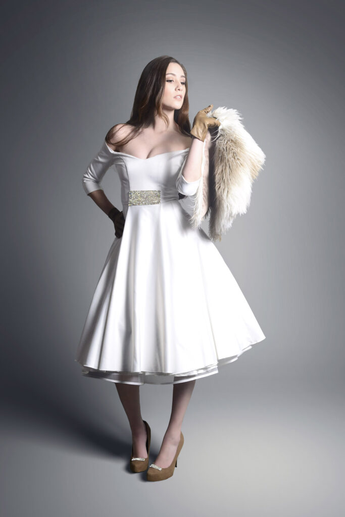 Melissa Lee is elegantly dressed in a white full-skirted dress with a fur accessory posing against a gradient gray backdrop.